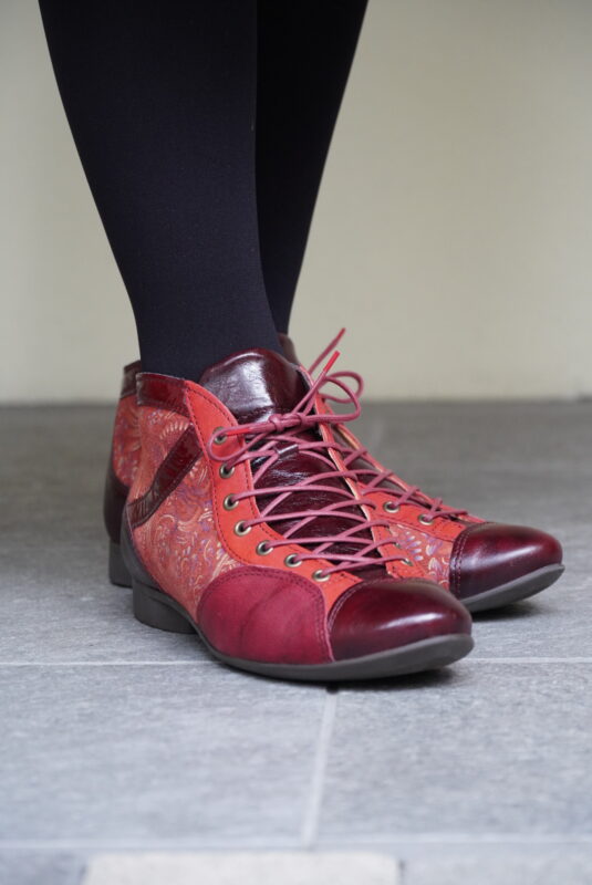 赤とワインレッドのレースアップ・アンクルブーツを履いた女性の足元。黒のタイツとくみあわせ、ペイズリー柄のパネルが入ったブーツが鮮やかに映えている。