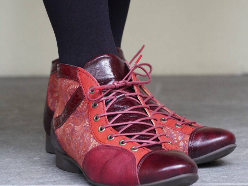 赤とワインレッドのレースアップ・アンクルブーツを履いた女性の足元。黒のタイツとくみあわせ、ペイズリー柄のパネルが入ったブーツが鮮やかに映えている。