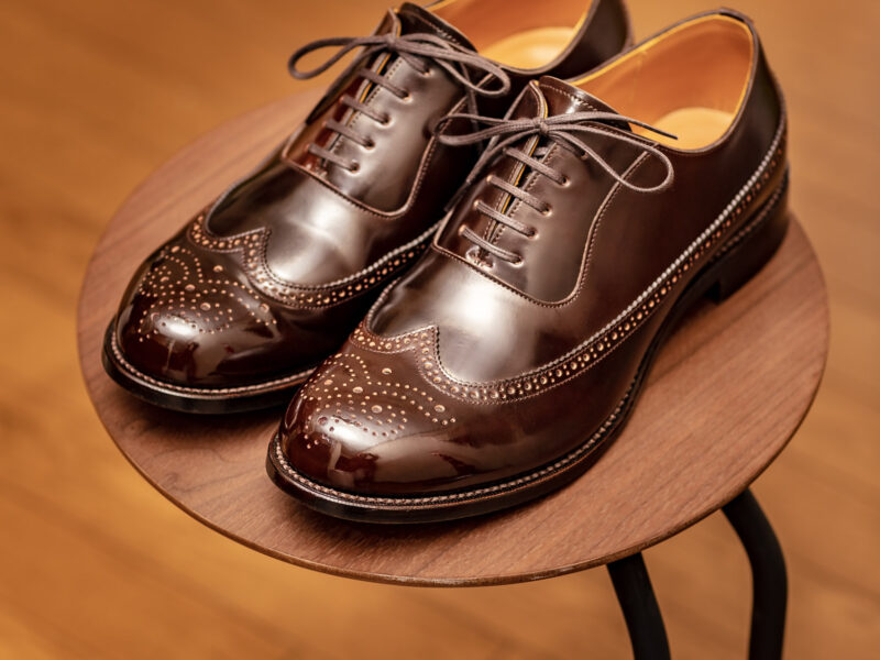 深いダークコニャック・コードバンのオーダーシューズ。温かみのある木の床と丸テーブルに置かれ、上質な革の艶と精密なメダリオンが際立っています。